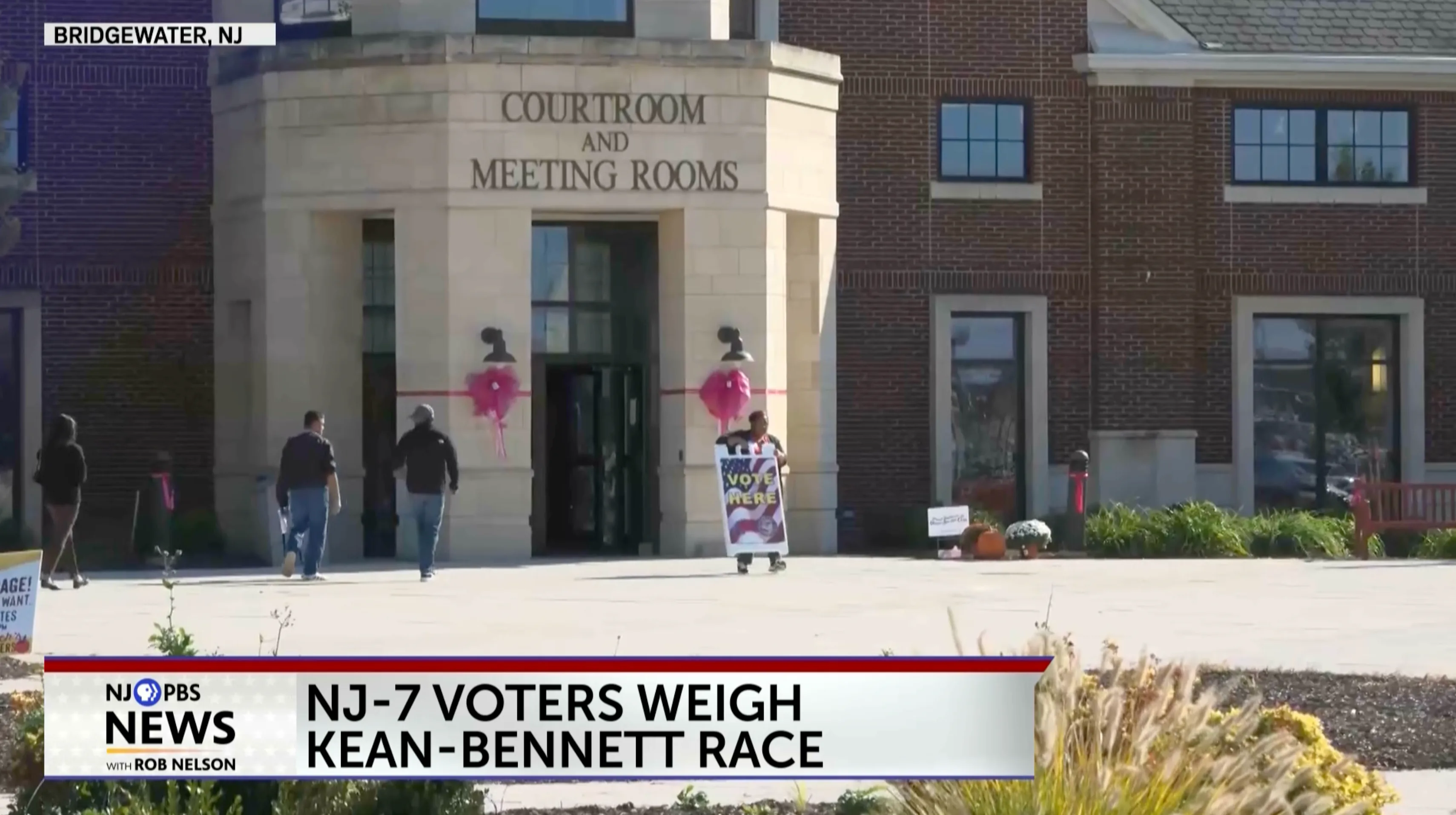an image of an exterior of a courthouse that is being prepared to serve as a polling station for voters. a woman is carrying a VOTE HERE sign.