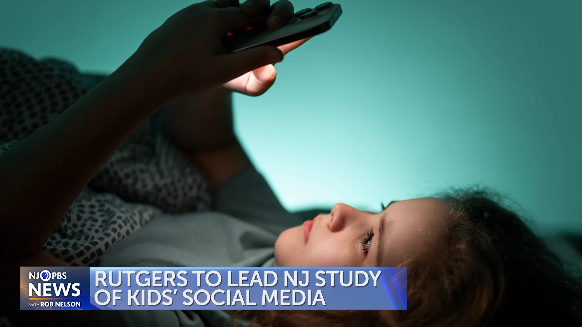 A child lies in bed looking at a smartphone in a dimly lit room. A lower-third graphic reads, “Rutgers to lead NJ study of kids’ social media,” with NJ PBS News branding.