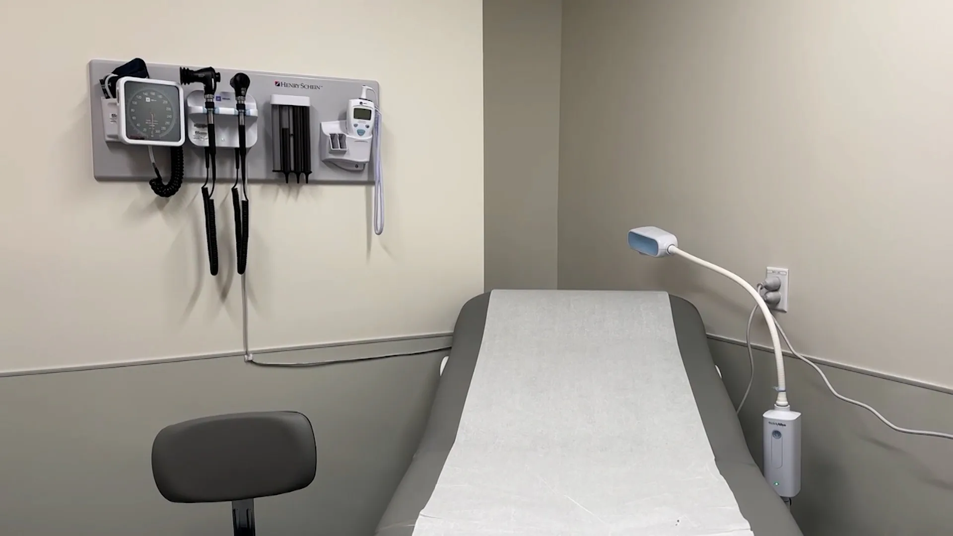 An empty examination table beside a chair, examination lamp and wall-mounted medical instruments.
